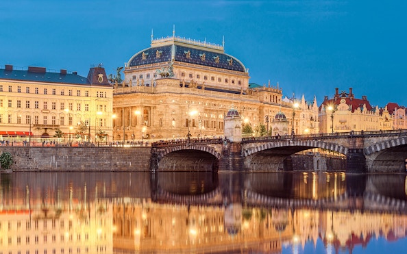 Prague's National Theatre and bridge illuminated at night, viewed from a river.