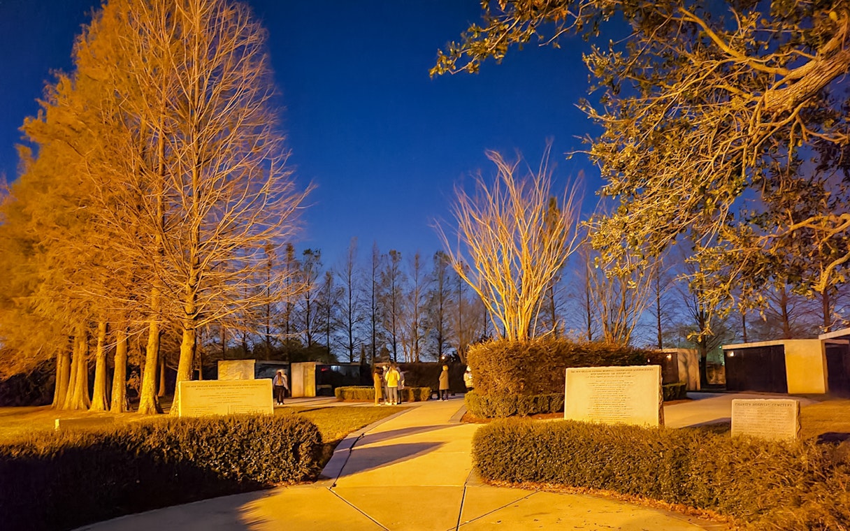 Nighttime view of a New Orleans cemetery with illuminated trees and visitors exploring pathways.