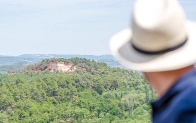 View of lush green hills near Moustier Sainte Marie, Provence, with a person in a hat in the foreground.