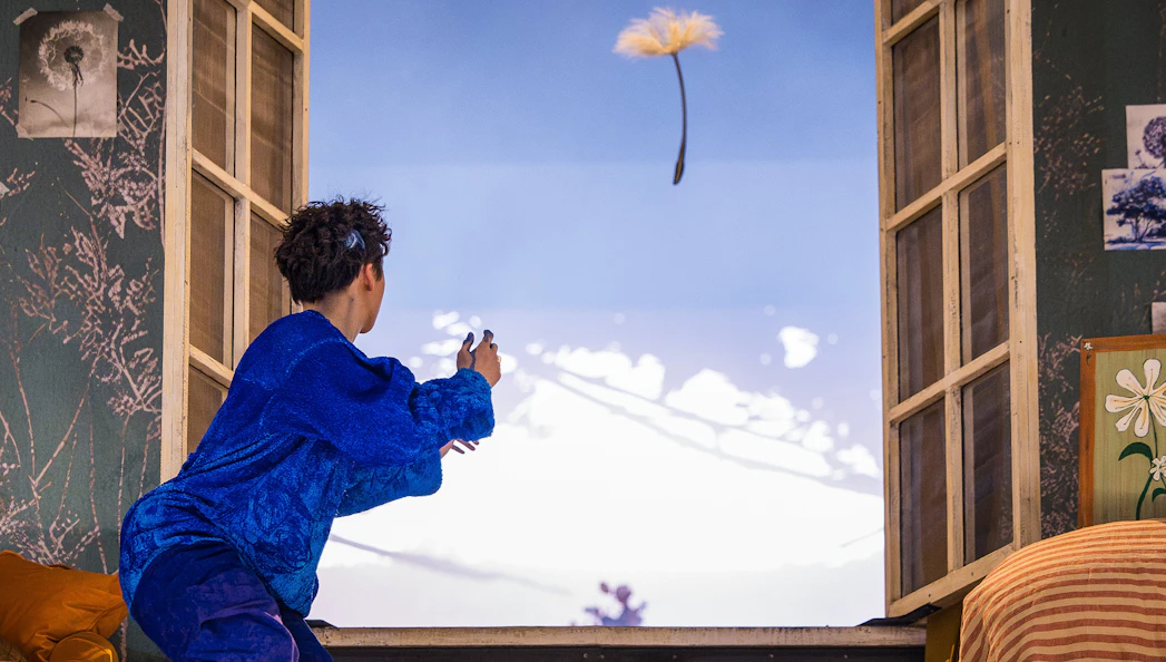 Performer in blue costume reaching towards floating dandelion during Cirque du Soleil Alizé show.