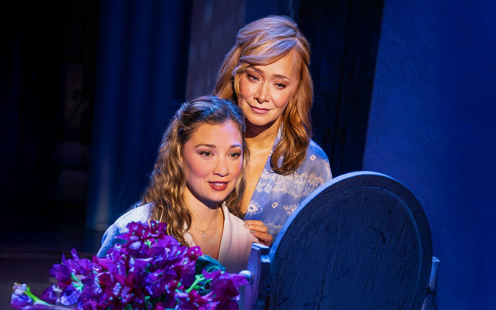 Two women in a scene from the musical Mamma Mia! with purple flowers in the foreground.