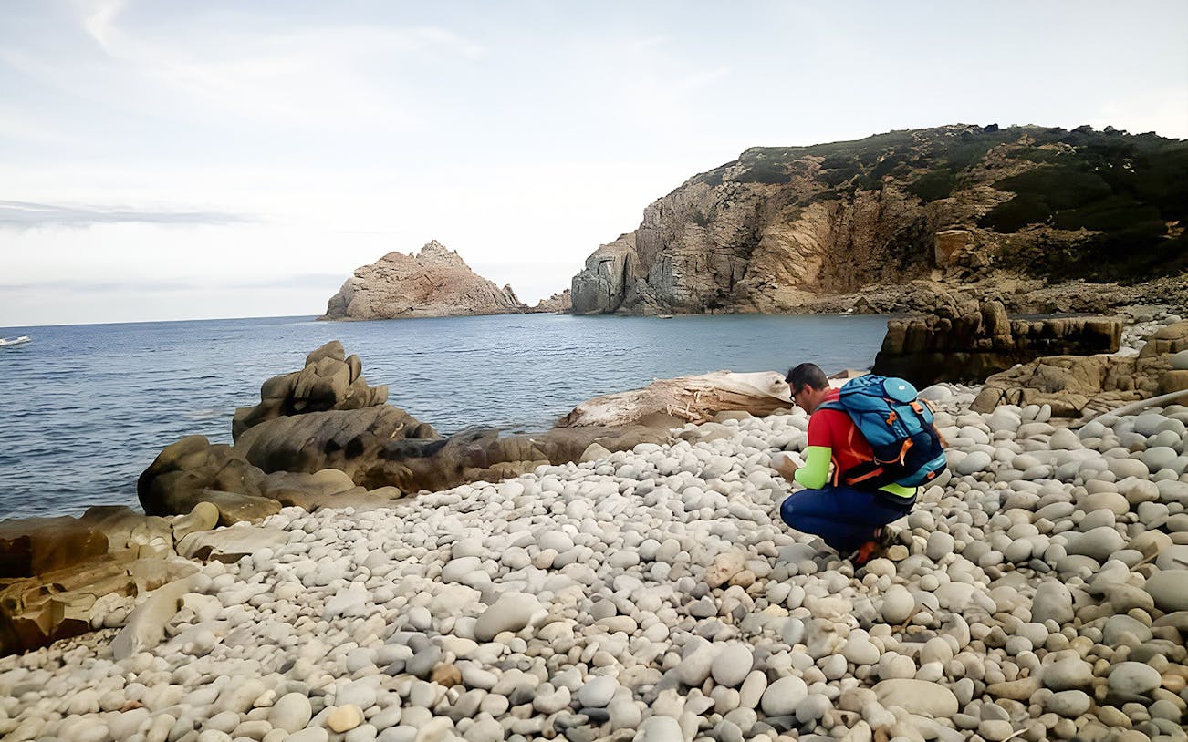 Trekker exploring rocky shore at Capo Pecora, Sardinia, with sea and cliffs in background.