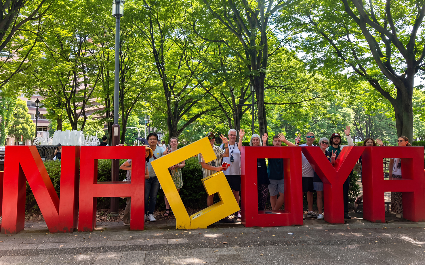Tourists posing with Nagoya sign in a park, promoting the Nagoya Grand Sumo Tournament.
