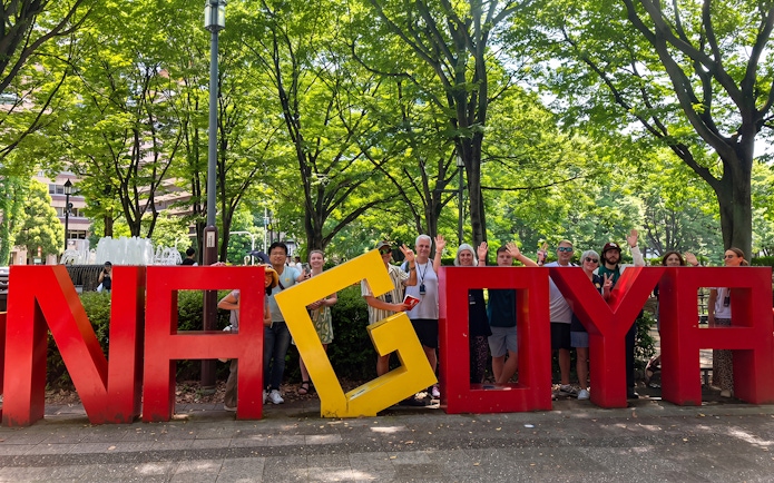 Tourists posing with Nagoya sign in a park, promoting the Nagoya Grand Sumo Tournament.