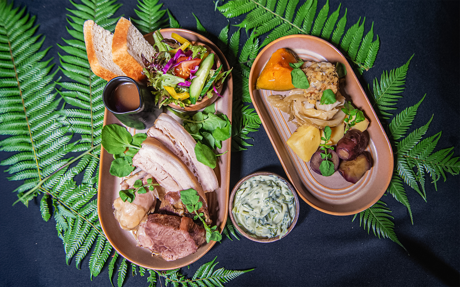 Two plated Hāngī meal with meats, vegetables, salad, and sauce on fern leaves.