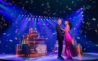 Actors performing in "Two Strangers (Carry a Cake Across New York)" on a Broadway stage with suitcases.