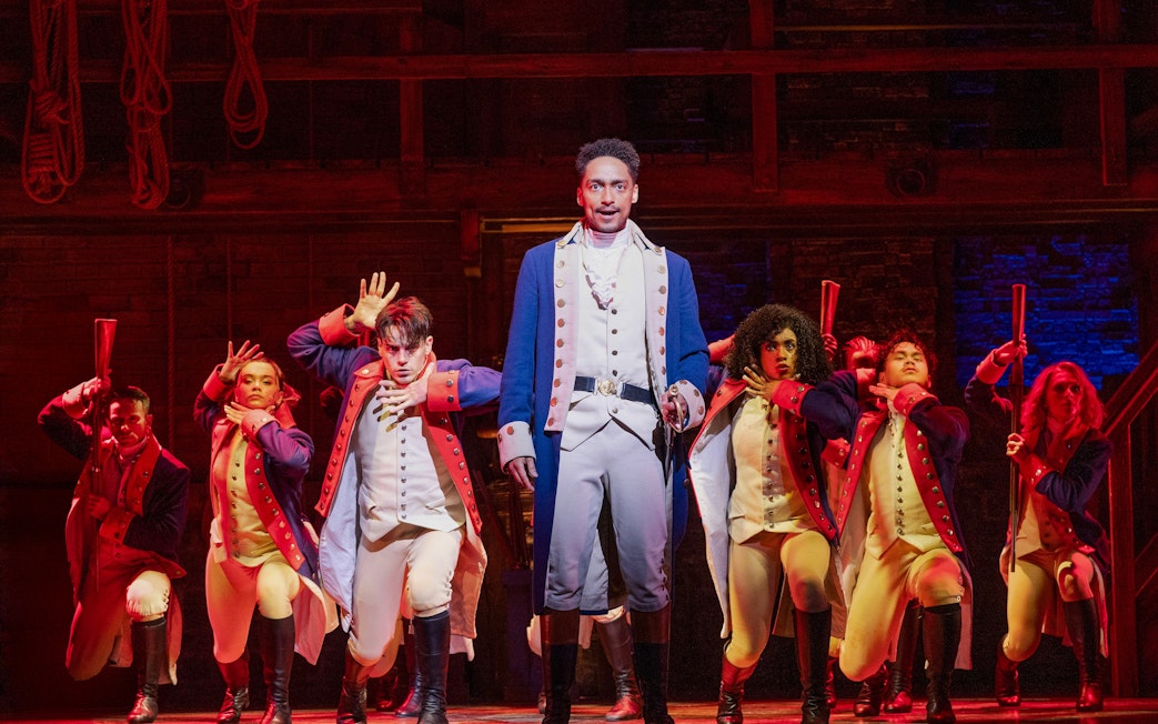 Performers in colonial costumes on stage during a Hamilton musical scene.