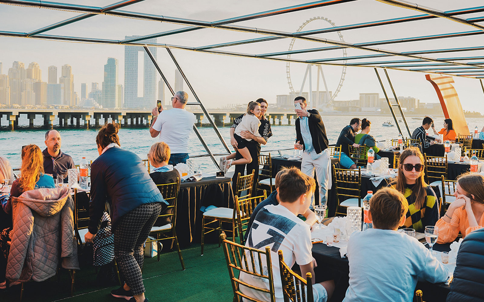 Guests dining on a sunset cruise in Dubai Marina with city skyline and Ferris wheel in view.