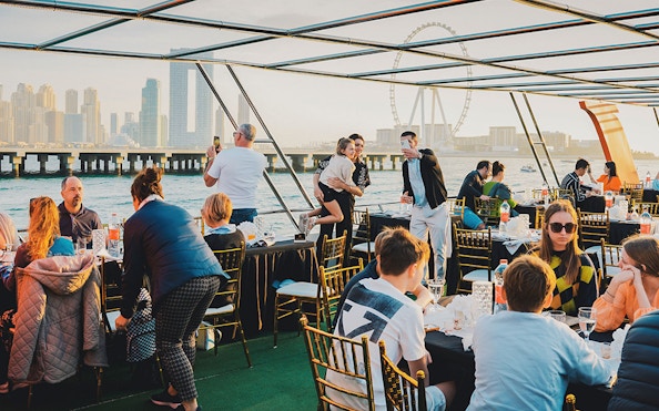 Guests dining on a sunset cruise in Dubai Marina with city skyline and Ferris wheel in view.