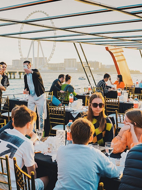 Guests dining on a sunset cruise in Dubai Marina with city skyline and Ferris wheel in view.