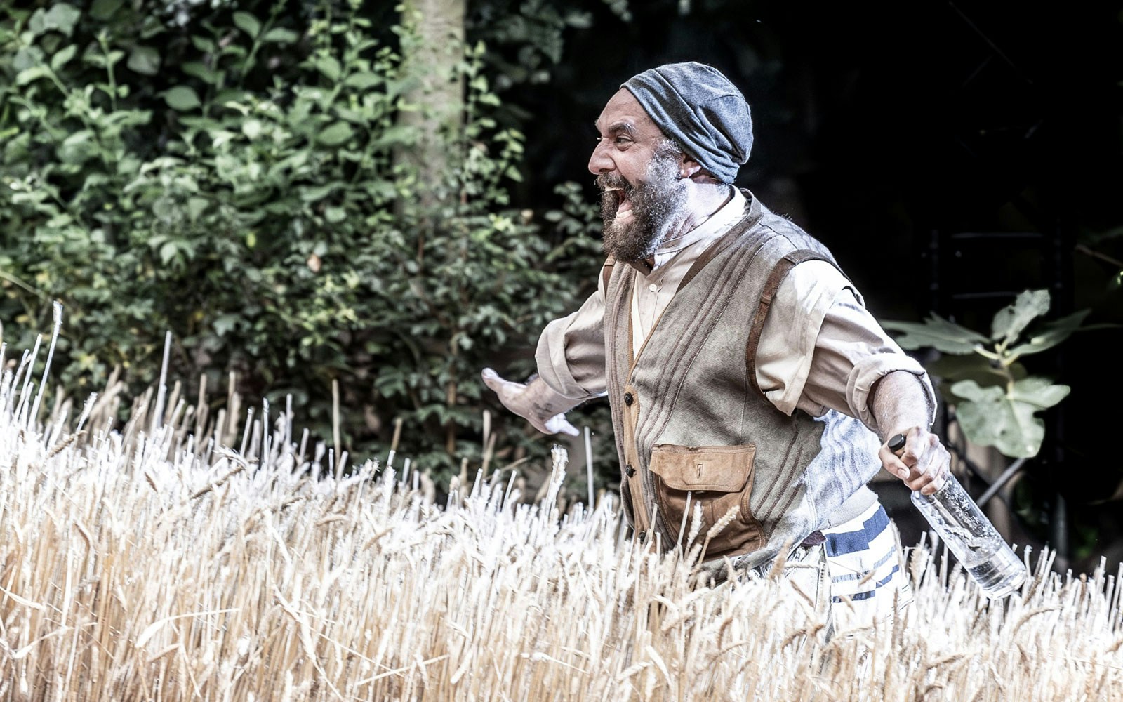 Man in traditional attire holding a bottle, performing in a scene from "Fiddler on the Roof" outdoors.