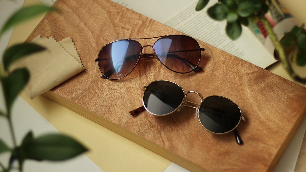 Two pairs of sunglasses on a wooden surface with a cleaning cloth nearby.