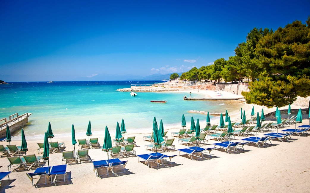Sun loungers and umbrellas on Ksamil beach, Albania, with turquoise water and trees in the background.