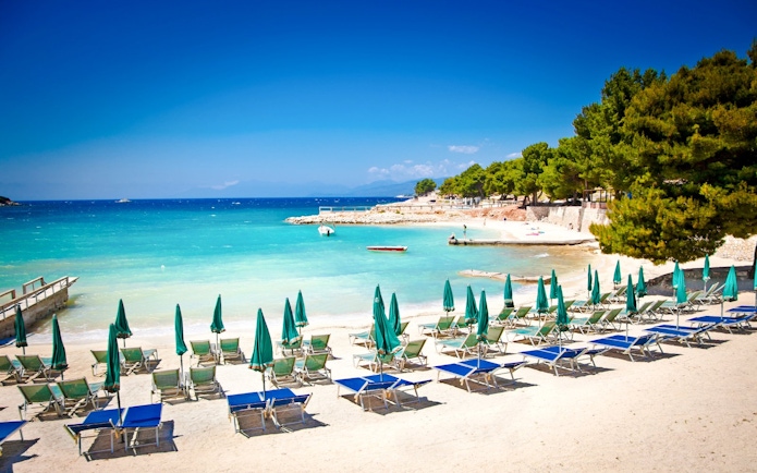 Sun loungers and umbrellas on Ksamil beach, Albania, with turquoise water and trees in the background.