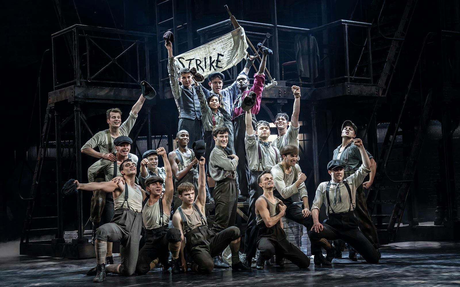 Performers in period costumes holding a "Strike" banner during a Newsies musical scene.