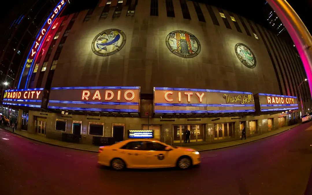Radio City Music Hall at night, New York, with a yellow taxi passing by.