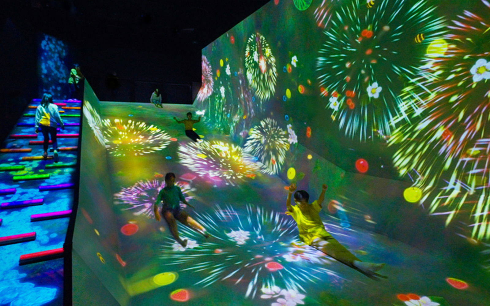Children sliding down a colorful, digitally projected slope with fireworks in a dark room.