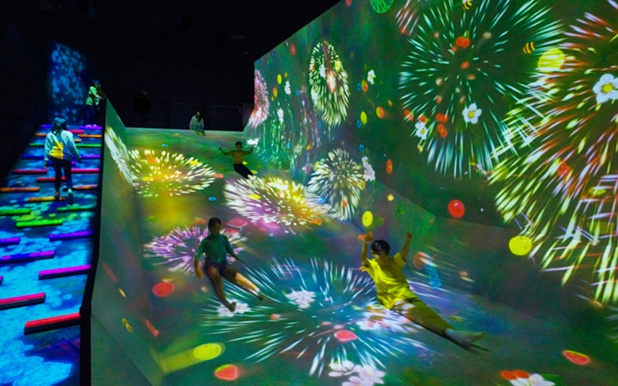 Children sliding down a colorful, digitally projected slope with fireworks in a dark room.
