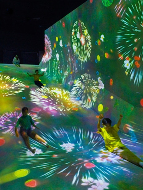 Children sliding down a colorful, digitally projected slope with fireworks in a dark room.