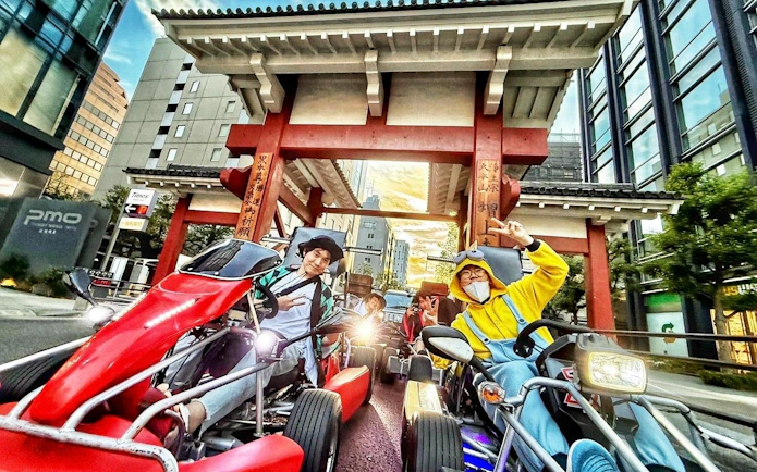 Friends in costumes on go-karts at Tokyo Tower tour, posing under a traditional gate.