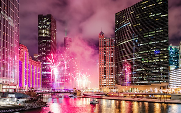 Fireworks display over the Chicago River at Wolf Point, surrounded by illuminated skyscrapers.