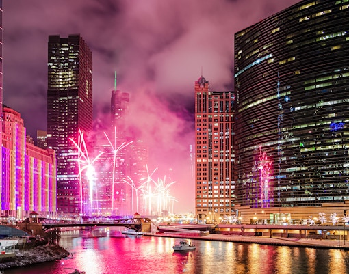 Fireworks display over the Chicago River at Wolf Point, surrounded by illuminated skyscrapers.