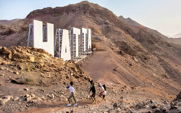 Hikers ascending rocky terrain near large Hatta sign during guided tour.