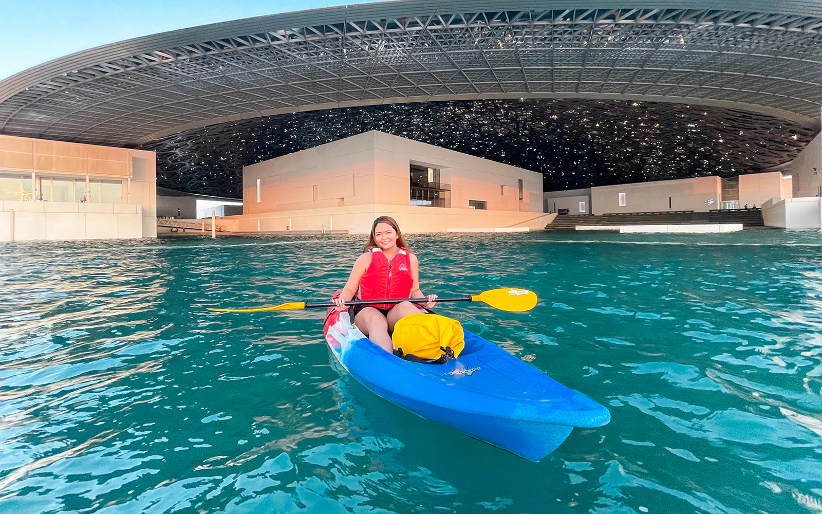 A kayaker in a blue kayak paddles beneath the Louvre Abu Dhabi dome.