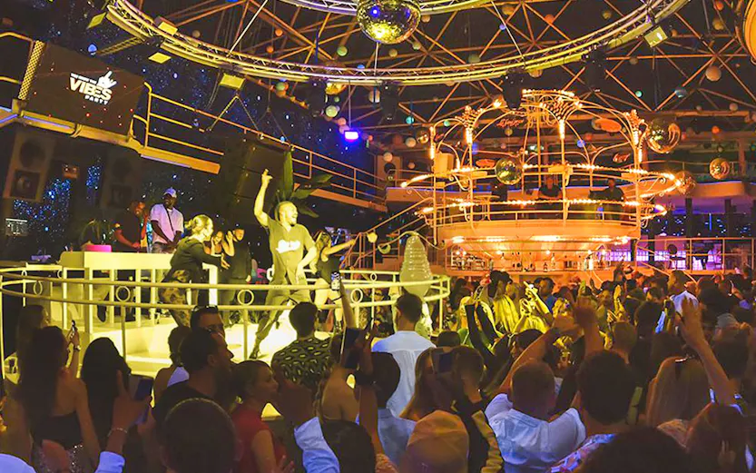 Crowded dance floor at Vibes Party, Es Paradis, with vibrant lighting and DJ booth.