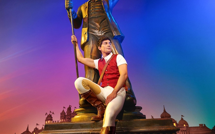 Man in red vest sitting by a large statue with a colorful sky backdrop, related to Wicked.