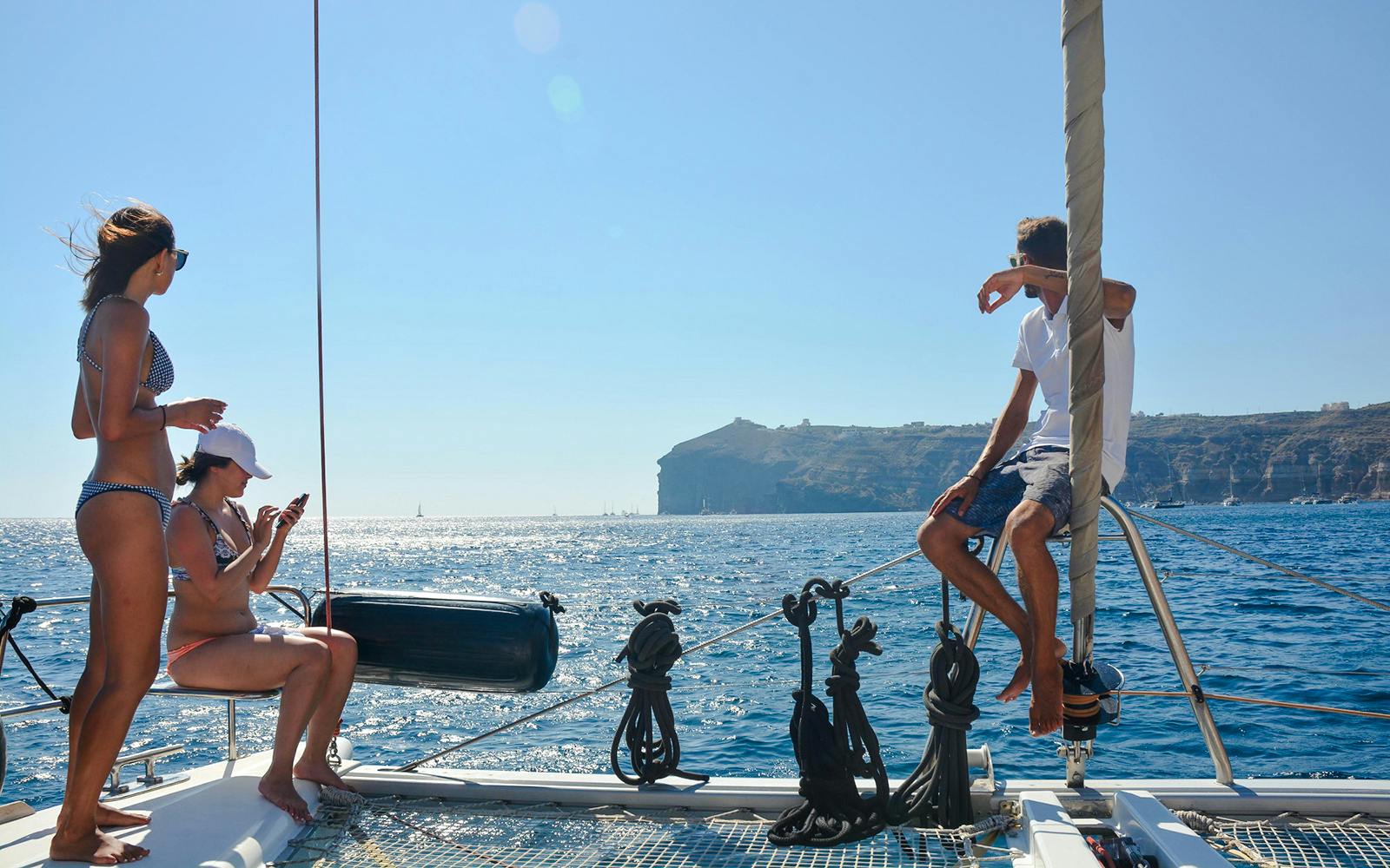 Guests relaxing on a catamaran with Santorini cliffs in the background during daytime cruise.