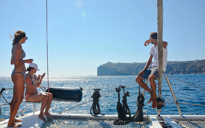 Guests relaxing on a catamaran with Santorini cliffs in the background during daytime cruise.