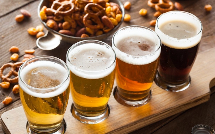Four glasses of beer on a wooden flight board with pretzels and nuts.
