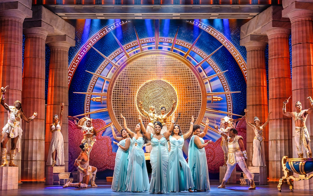 Stage performance of Disney's Hercules with actors in Greek costumes and ornate backdrop.