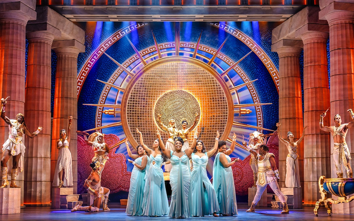 Stage performance of Disney's Hercules with actors in Greek costumes and ornate backdrop.