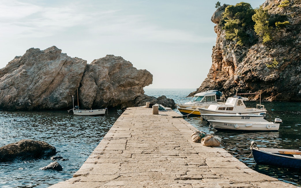 Stone pier with boats in Dubrovnik West Harbour, Croatia, surrounded by rocky cliffs.