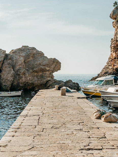 Stone pier with boats in Dubrovnik West Harbour, Croatia, surrounded by rocky cliffs.