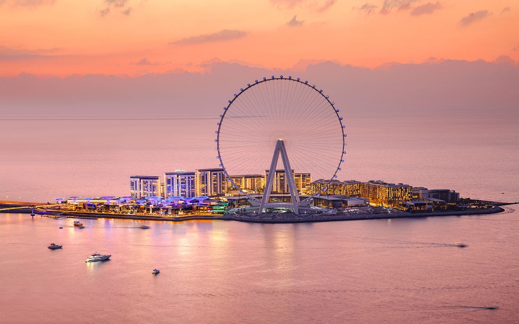 Ain Dubai Ferris wheel at sunset with illuminated cityscape and boats on the water.