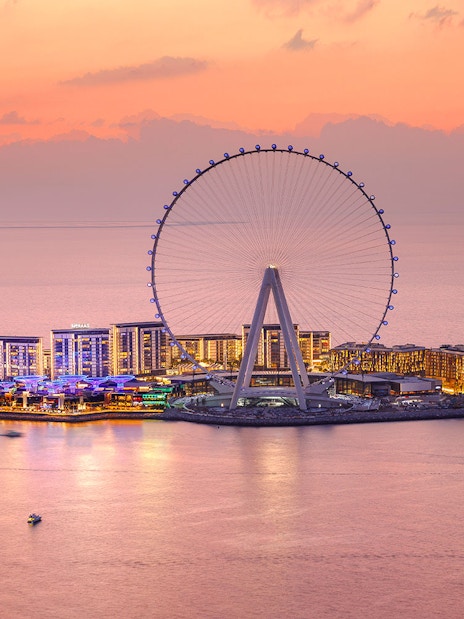 Ain Dubai Ferris wheel at sunset with illuminated cityscape and boats on the water.