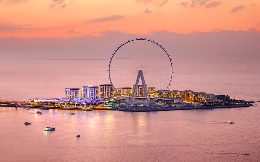 Ain Dubai Ferris wheel at sunset with illuminated cityscape and boats on the water.