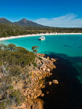Catamaran in turquoise waters of Wineglass Bay, Tasmania, with forested hills and rocky shoreline.