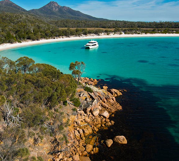 Catamaran in turquoise waters of Wineglass Bay, Tasmania, with forested hills and rocky shoreline.