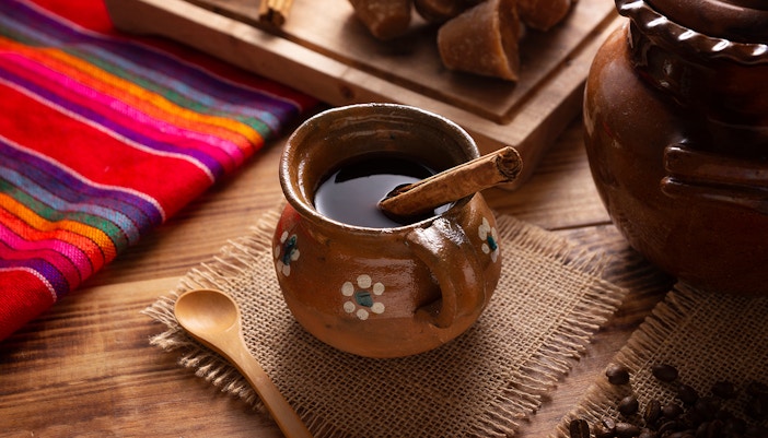 Cafe de Olla in a jarrito with coffee beans, cinnamon sticks, and piloncillo on rustic wood surface.