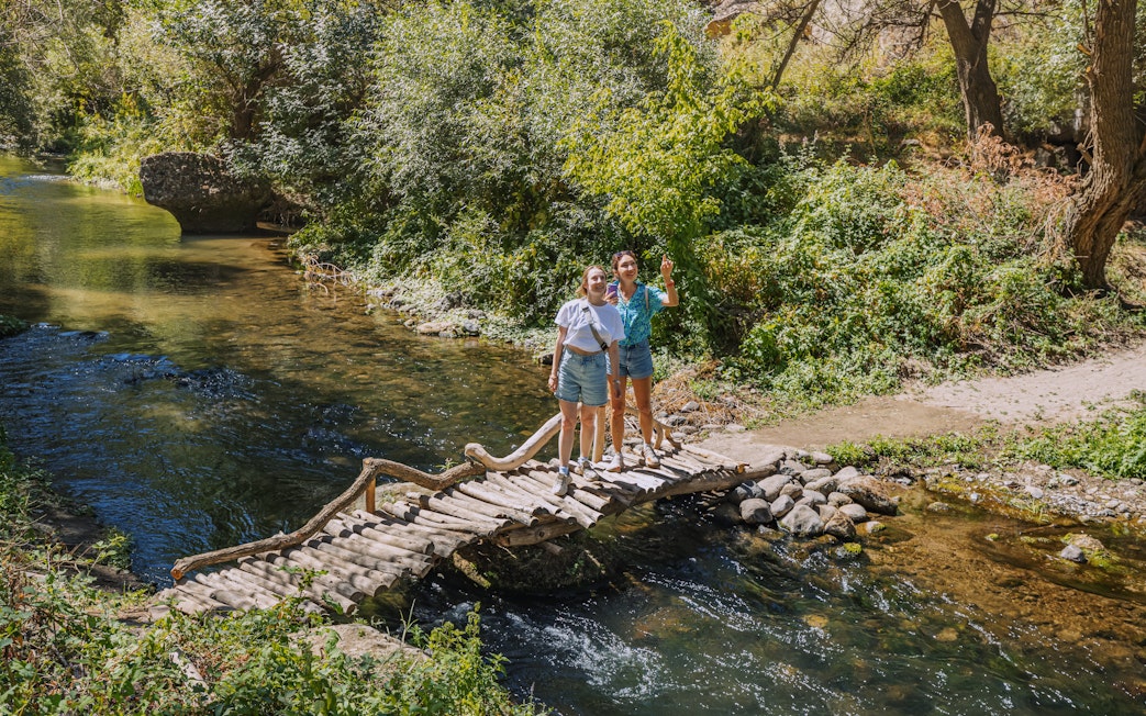 Tourists on a wooden footbridge over the Melendiz River surrounded by lush greenery.