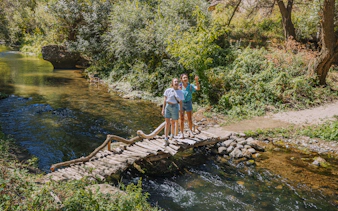 Tourists on a wooden footbridge over the Melendiz River surrounded by lush greenery.