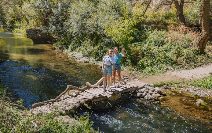 Tourists on a wooden footbridge over the Melendiz River surrounded by lush greenery.