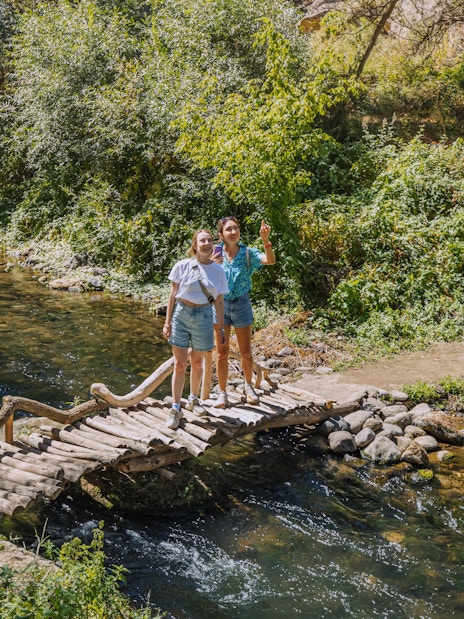 Tourists on a wooden footbridge over the Melendiz River surrounded by lush greenery.