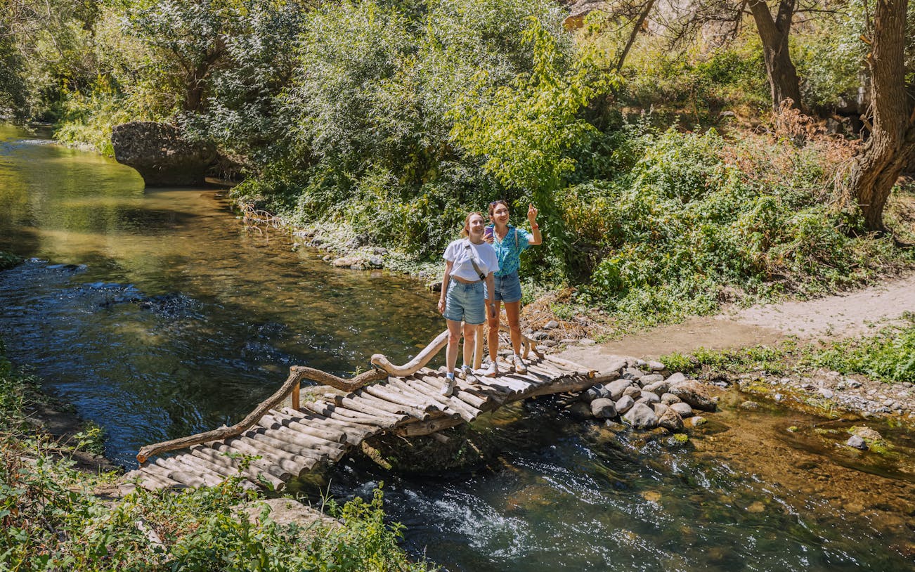 Tourists on a wooden footbridge over the Melendiz River surrounded by lush greenery.