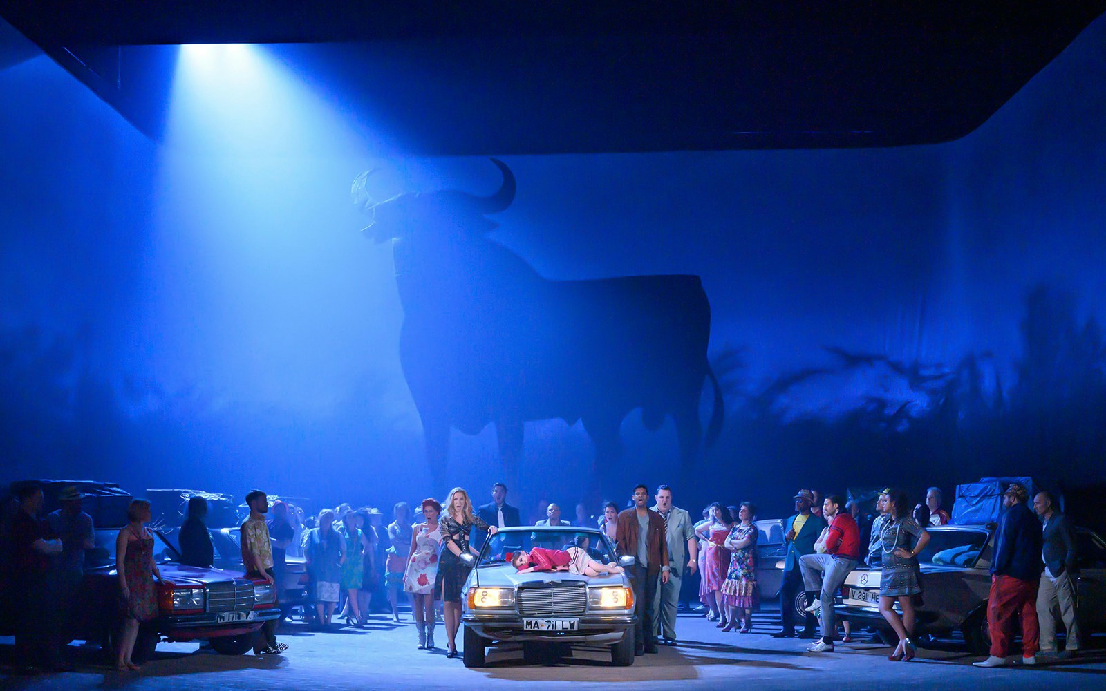 Opera scene with performers around a car under a bull silhouette, related to Carmen.