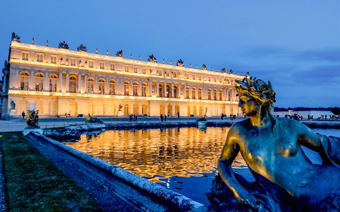 Versailles Palace illuminated at night with fountain statue in foreground, Paris tour.